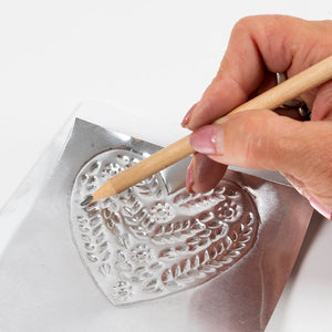 Hand using a wooden pencil to press intricate designs into a metal mold on a white background