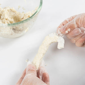 Hands wearing gloves stretching dough on a white surface with a glass bowl of dough in the background.