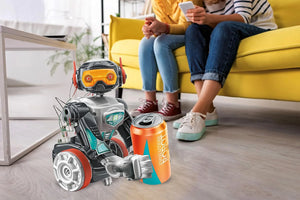 Toy robot holding a can in a living room with two people sitting on a yellow couch.