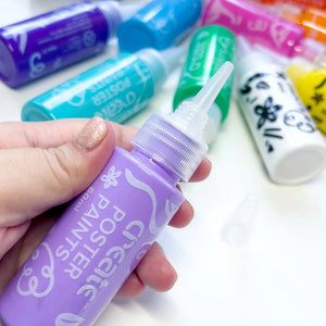 a hand holding a purple poster paint tube with a nozzle top with other paints on a white table in the background