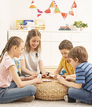 Four children sitting around a woven table playing cards in a room with colorful decorations.