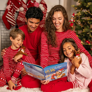 Family of four in matching red pajamas reading a book in front of a Christmas tree.