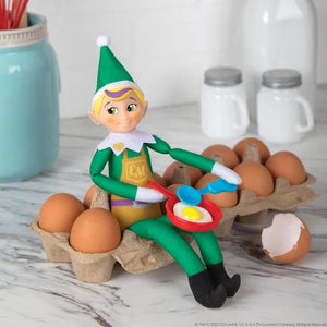 Elf figurine cooking an egg in a frying pan on a carton of eggs with kitchen items in the background.