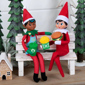 Two Elf on the Shelf dolls sitting on a bench with food items, surrounded by Christmas decorations.