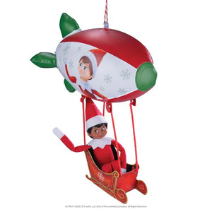 elf on the shelf sitting in a sleigh that's attached to a red and white blimp overhead on a white background