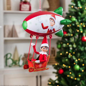 elf on the shelf sitting in a sleigh that's attached to a red and white blimp overhead with a christmas tree and shelving in the background