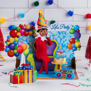 An elf scout sitting on a pop up chair with birthday decorations and presents in the background. Pop up is from The Elf on the Shelf 20th Anniversary - Pop Ups set