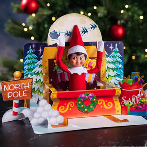 An elf scout sitting on a pop up Santa Sleigh with snow and tress in the background and Santa flying overhead. Pop up is from The Elf on the Shelf 20th Anniversary - Pop Ups set