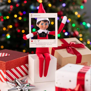 An elf on the shelf character sitting on presents posing with a cardboard cutout of an Instagram grid photo with a Christmas Tree in the background