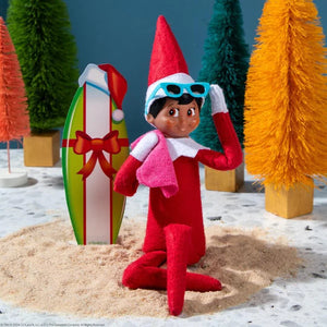 Elf on the Shelf character in a red outfit with sunglasses, sitting in sand with Christmas trees in the background.